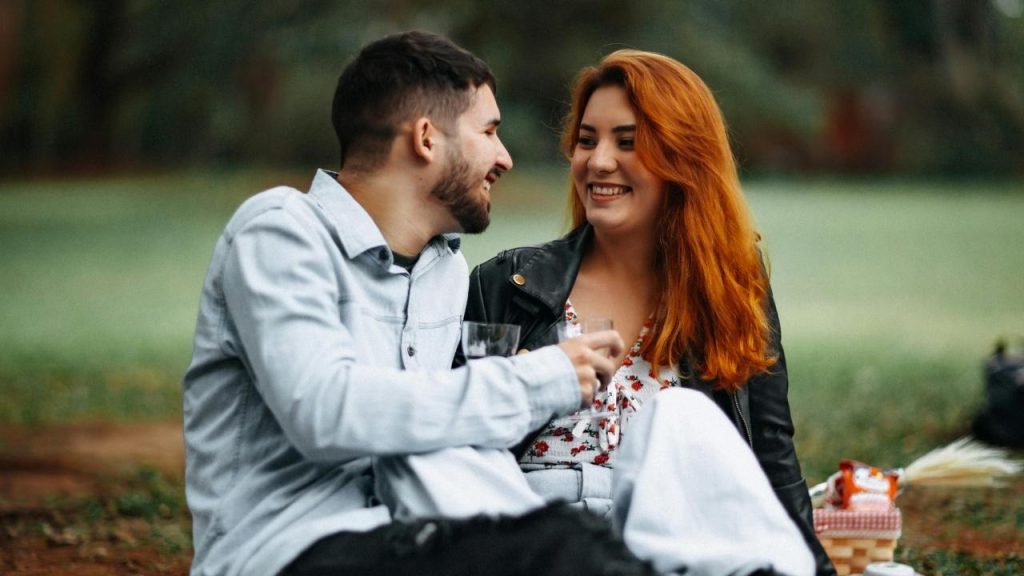 A man and a woman with red hair sit on the grass laughing during a picnic.