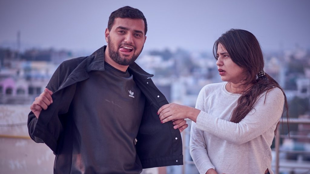 A man opening his jacket to show his shirt while standing outdoors on a rooftop, as a woman beside him looks at him with a concerned or surprised expression.