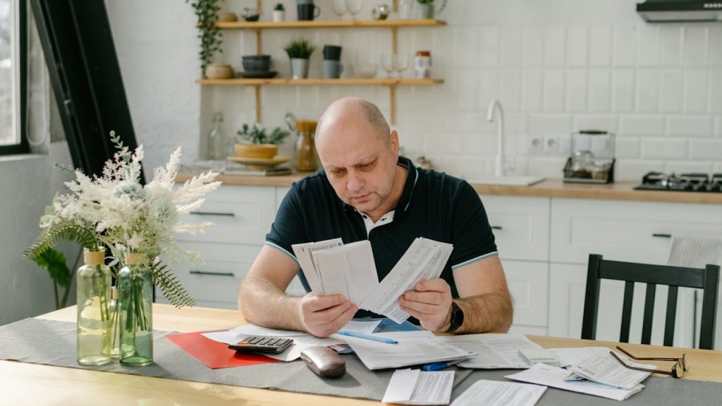A man appears to be worried, sitting at a table with papers and a calculator.
