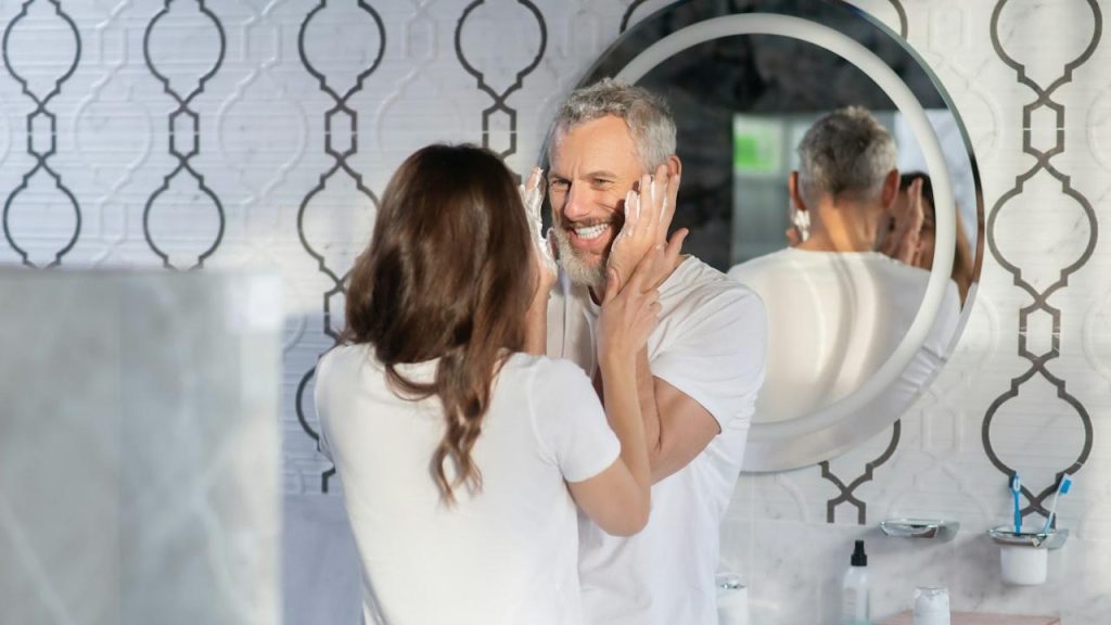 A woman holding a man’s hands as he smiles at her and tries to apply shaving foam to his beard as they stand in a bathroom.