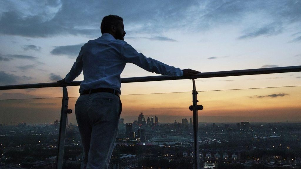 A man in a dress shirt and pants stands on a balcony railing at sunset, looking out at the city skyline.