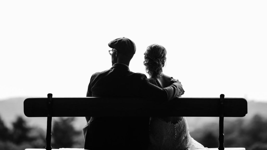 An older couple sitting on a bench with the man resting his hand on the woman’s shoulder.