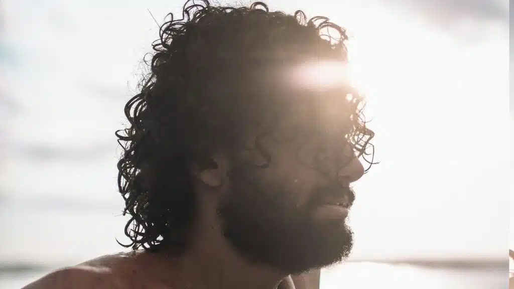 A bearded man looking into the distance while the sun shines behind him over a body of water.