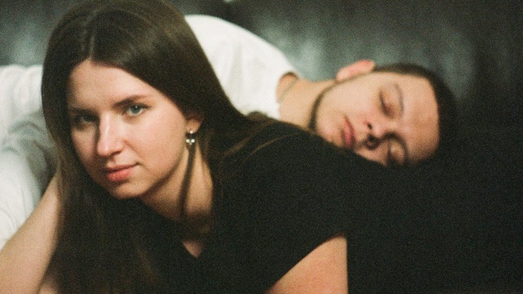 A woman lying on a couch with her sleeping boyfriend.