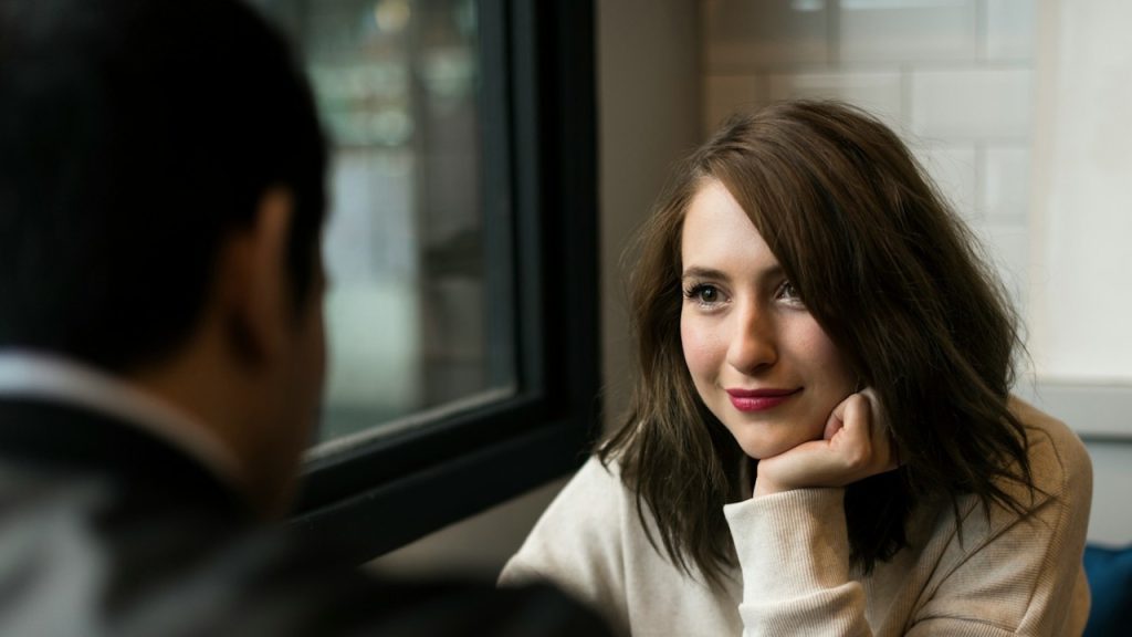 A woman smiling and staring at a man while holding her chin with her hand.