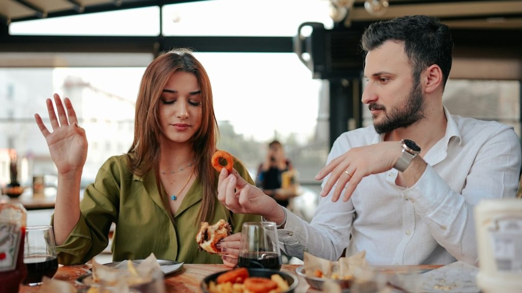 A man offering donuts to his girlfriend while they are having a meal in a restaurant.