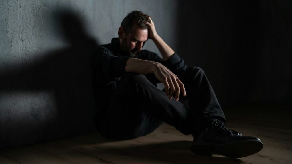 A man sits alone in a dark room with his hand on his head, looking stressed and sad.
