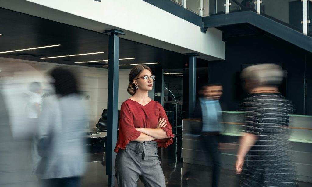 A woman wearing glasses standing in the midst of a crowd in an office with crossed arms.