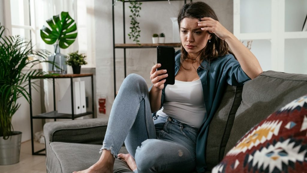 A frustrated woman sitting on a couch and looking at a phone’s screen.