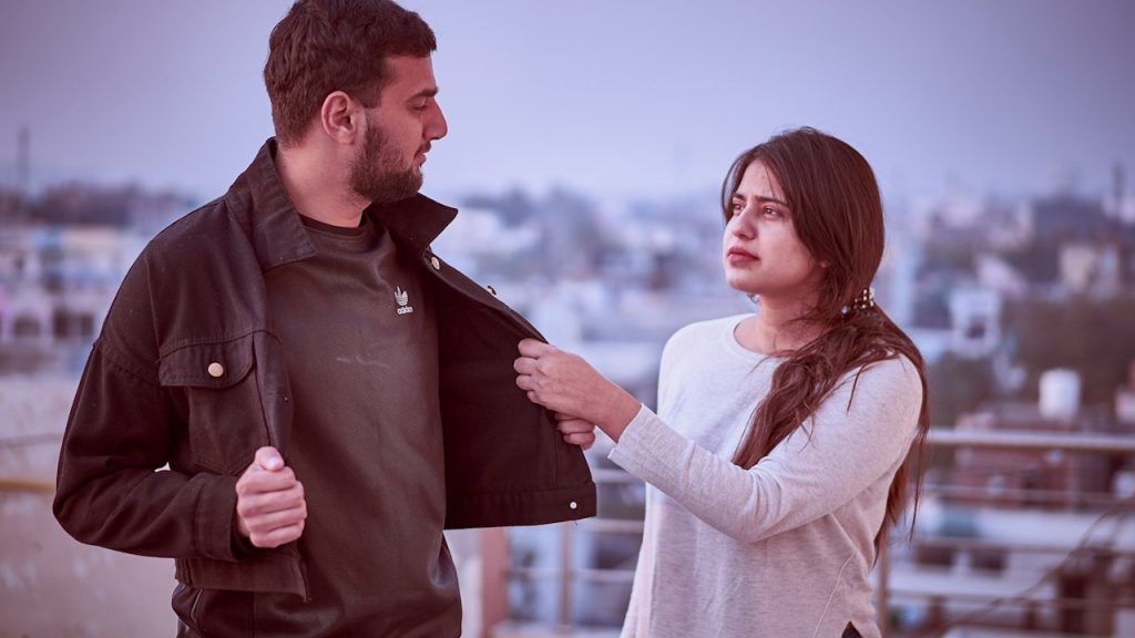 A woman tugging at a bearded man’s jacket and looking at him with a forlorn expression.