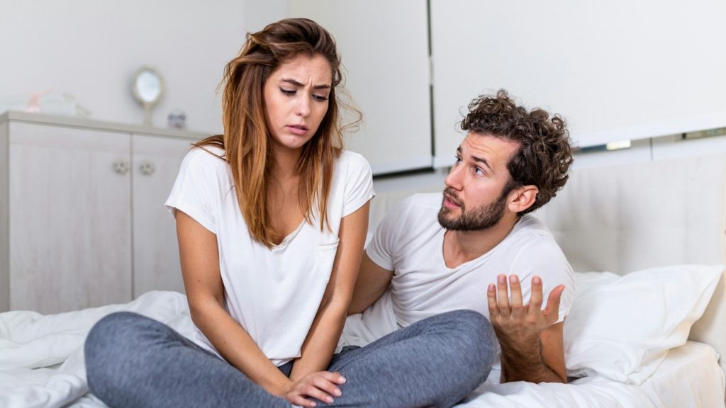 A man talking to her angry wife while they are sitting on a bed.