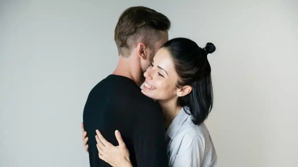 A smiling woman hugs a man from the front with her eyes closed, her face resting against his shoulder