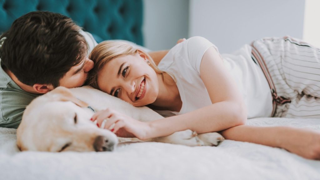 Couple lying in bed with their dog, with the man kissing the woman's forehead as she smiles.