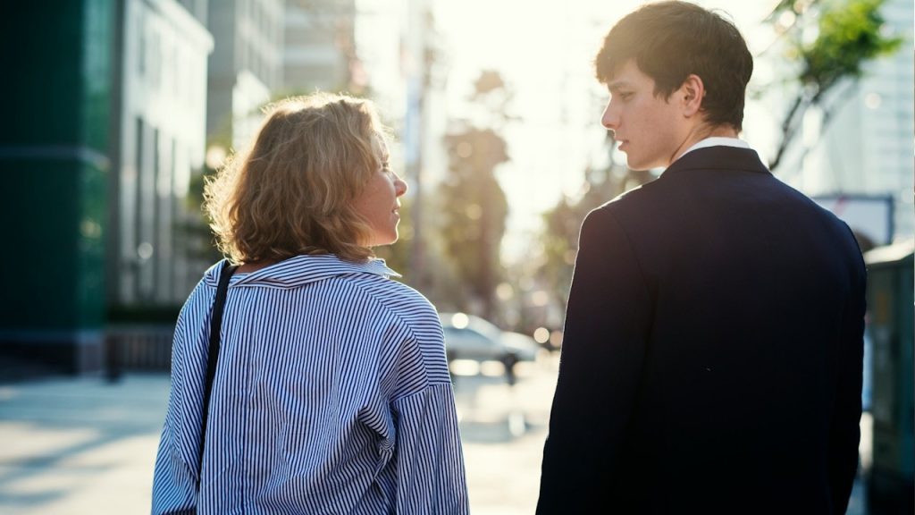 Woman in a striped shirt looking back at a man in a dark suit walking on a sunny city street.