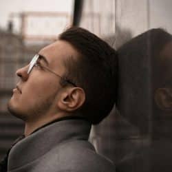 A serious man with glasses and a beard is leaning his head against a stone wall, looking up.