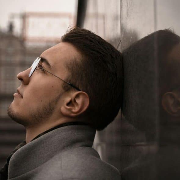 A serious man with glasses and a beard is leaning his head against a stone wall, looking up.