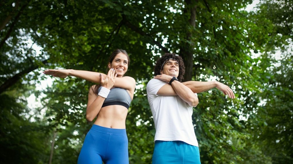Man and woman stretching their arms outdoors with a background of trees.