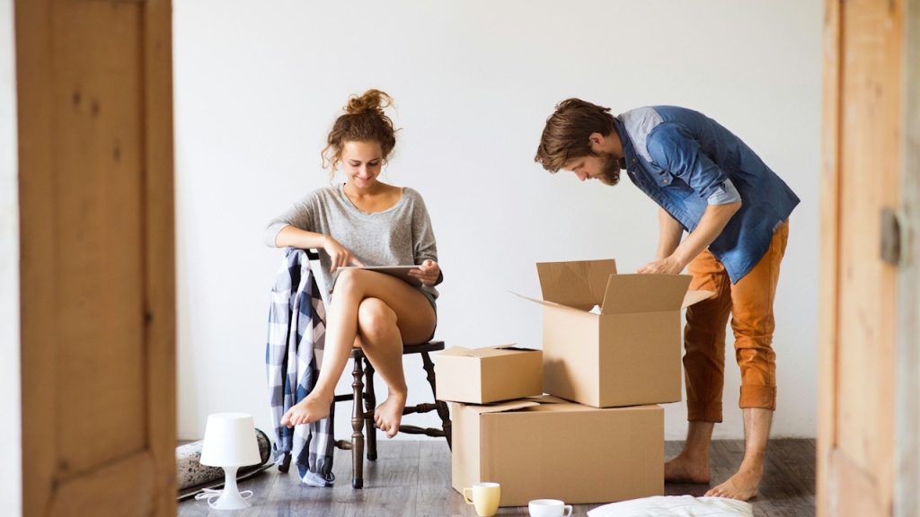 Couple moving into a new home, with the man unpacking cardboard boxes and the woman sitting on a chair looking at a tablet.