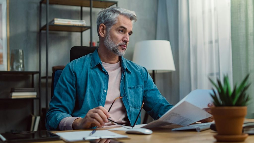A middle-aged bearded man tending to financial documents and looking perturbed.