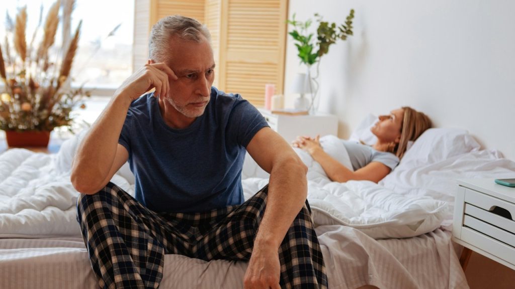 An older man sits on the edge of a bed looking distressed, while a woman lies in the background.