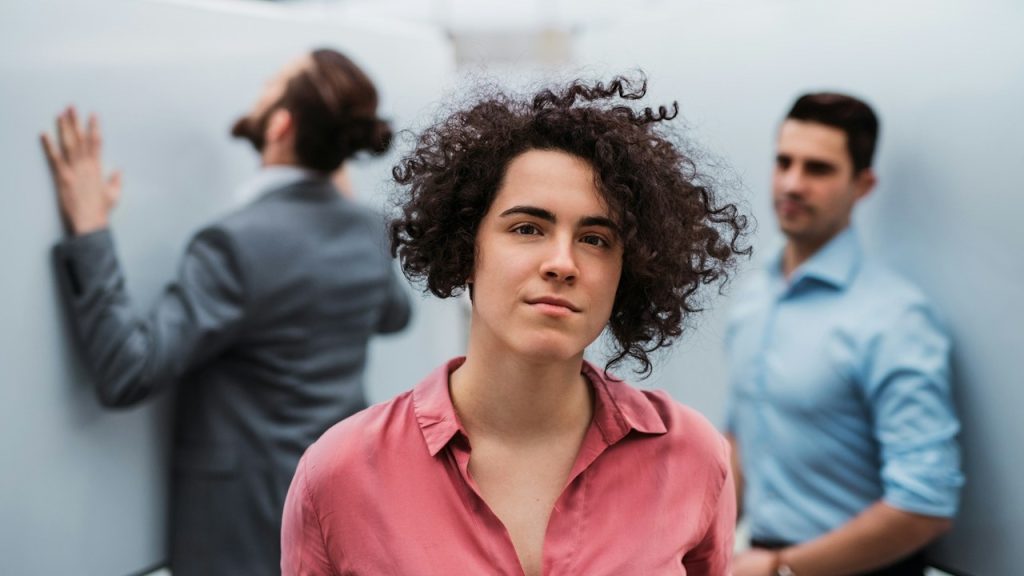 A woman with curly hair is looking at the camera, and two men are standing in the background.
