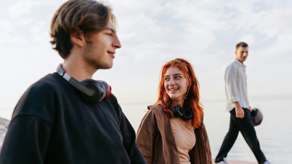 A couple smiling while wearing headphones and another man holding a helmet is passing by them.