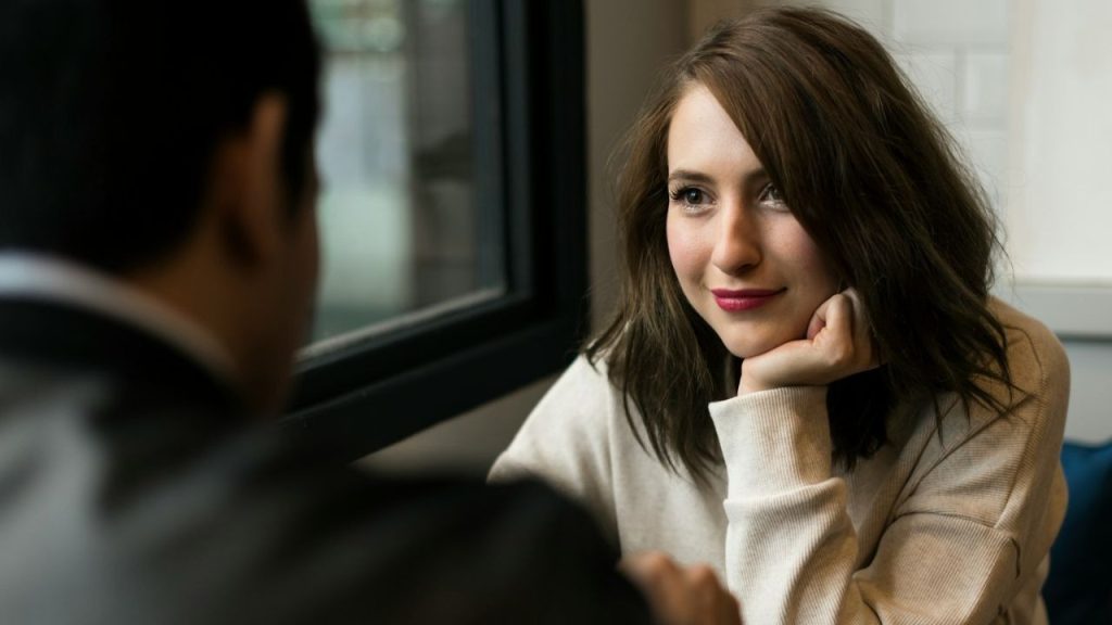 A woman with a slight smile sits across from a person, resting her chin on her hand.