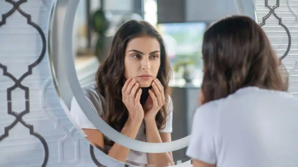 A woman with her hands on her face looks at her reflection in a round mirror.