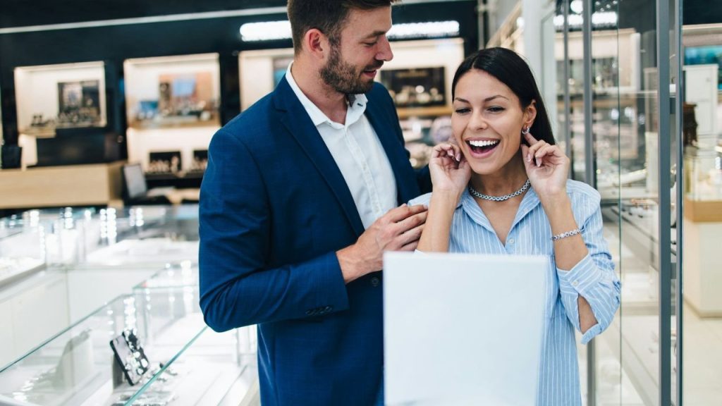 A couple is looking at jewelry in a store, and the woman is smiling excitedly.