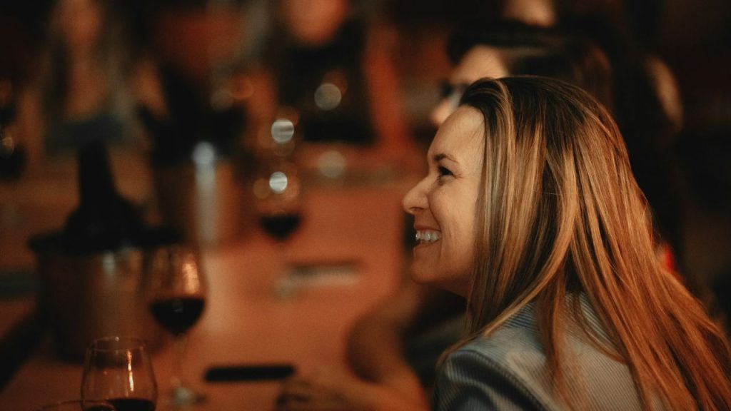 A woman with long, light brown hair smiles while looking to her right, with blurred wine glasses in the foreground.