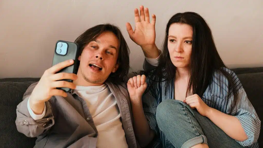 A man waving hands while making a video call and his wife is looking at the screen, seems annoyed.