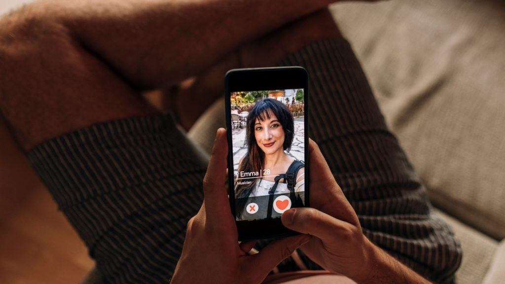 A person holding a smartphone showing a dating app profile of a woman, with options to like or reject the profile visible on the screen.