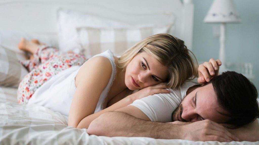 A pensive woman lies on her side, resting her head on a man's back, as they lie in bed.