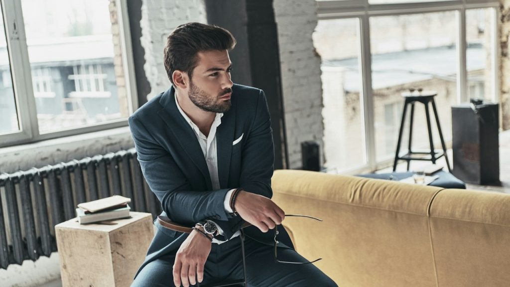 A bearded man wearing a suit and holding sunglasses in his hand sits on a chair in his living room.