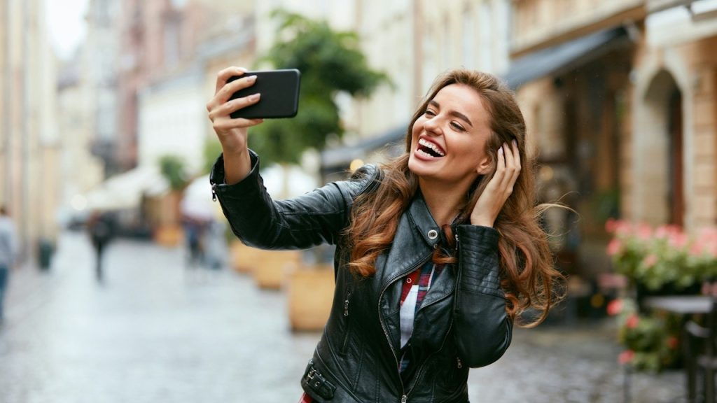 A woman smiling and taking a selfie while standing in a street and adjusting her hair.