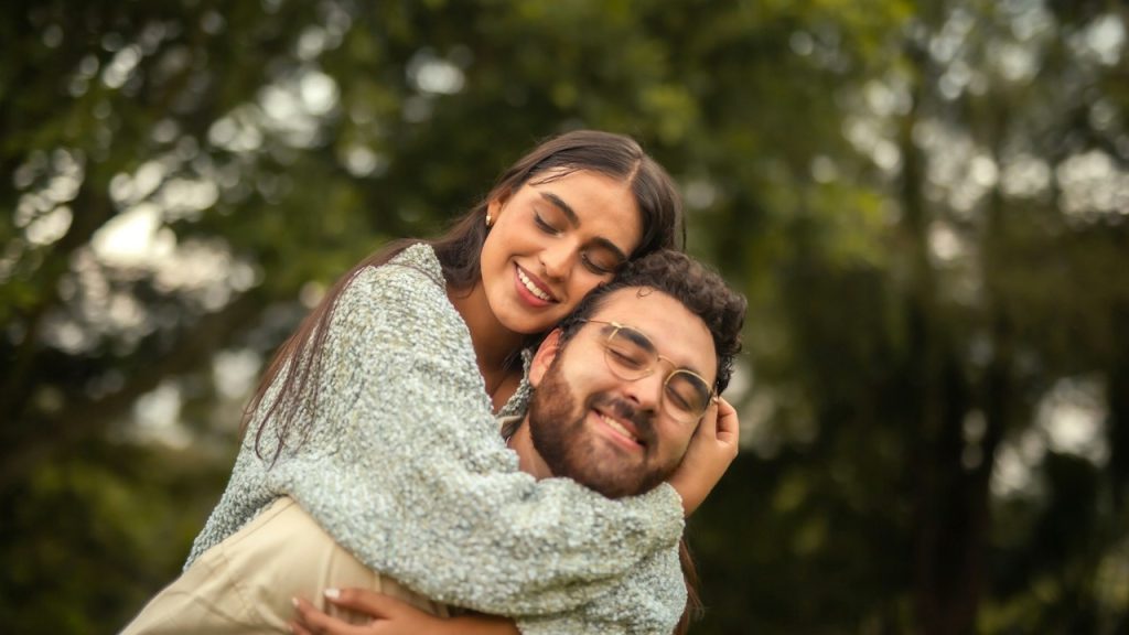 A bespectacled man carrying a woman on his back outdoors while they both have their eyes closed.