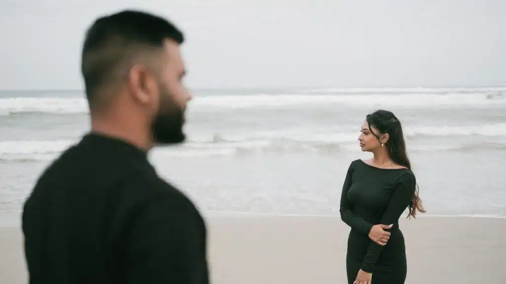 A man and a woman, both wearing black dresses, are standing at a beach.