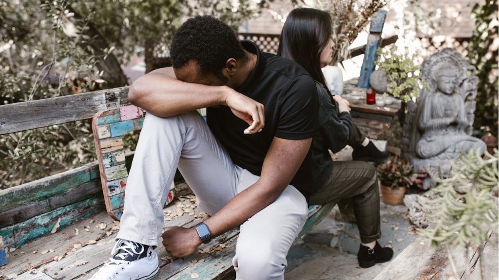 Black man with his head down on a bench, back-to-back with a woman outdoors.