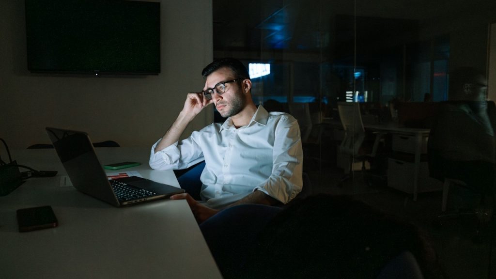 A bespectacled man looking at a laptop’s screen while holding his finger up to his temple and looking worried.