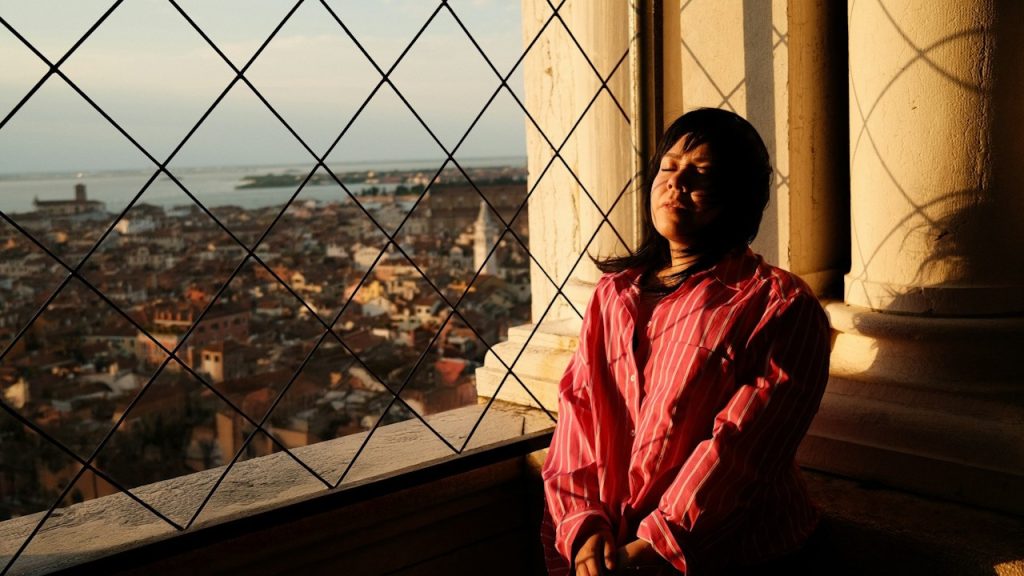 A depressed woman leaning on the wall near a window.