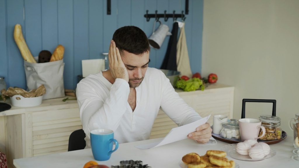 A worried man sits at a table, looking at some papers and holding his hand up to his temple.