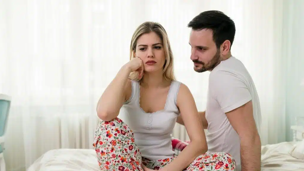 A sad woman is sitting on a bed, and her husband is looking at her while sitting next to her.