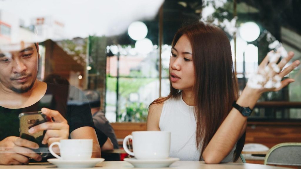 A man is looking at his phone while a woman sitting across from him at a cafe table is talking and gesturing with her hand