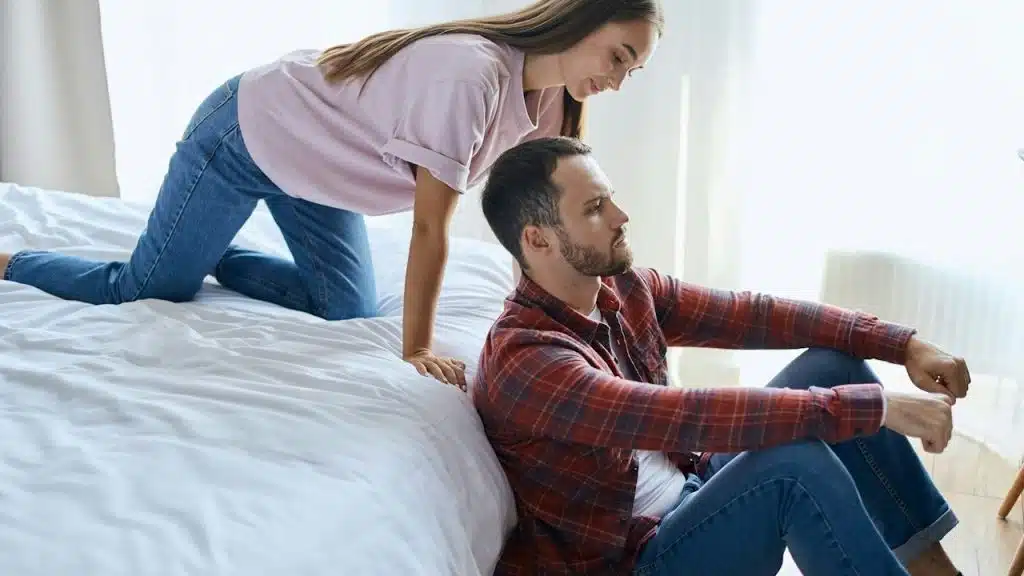 A man dressed in a casual outfit sits on a light-colored floor and listens to the news attentively. A pretty, playful woman creeping up from behind stands on the bed.