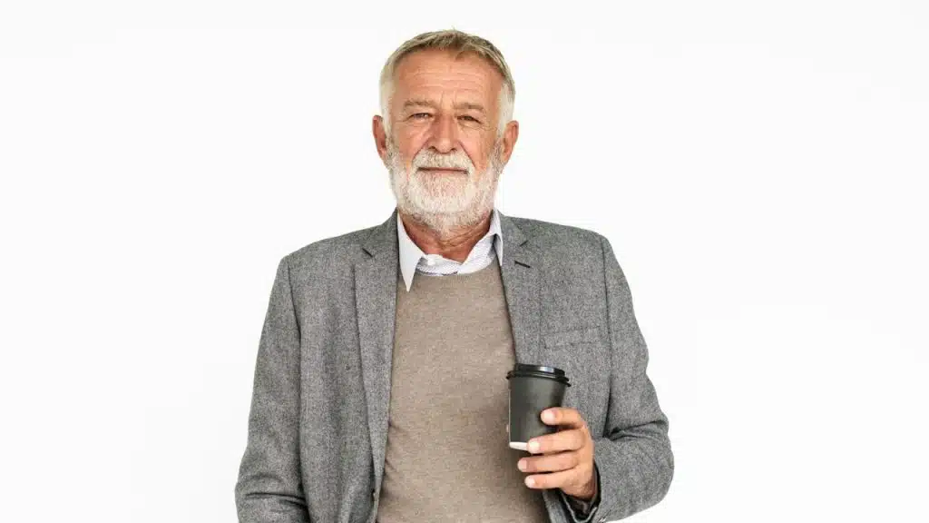 An old man wearing a suit who is holding a cup of coffee is standing against a white wall.