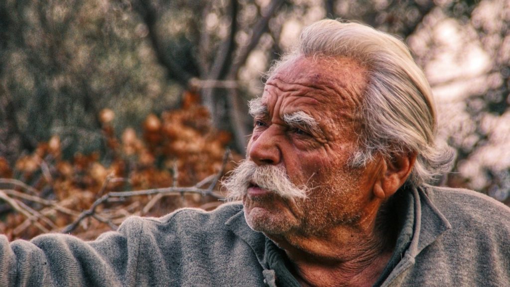 An old man wearing green shirt is standing in woods.