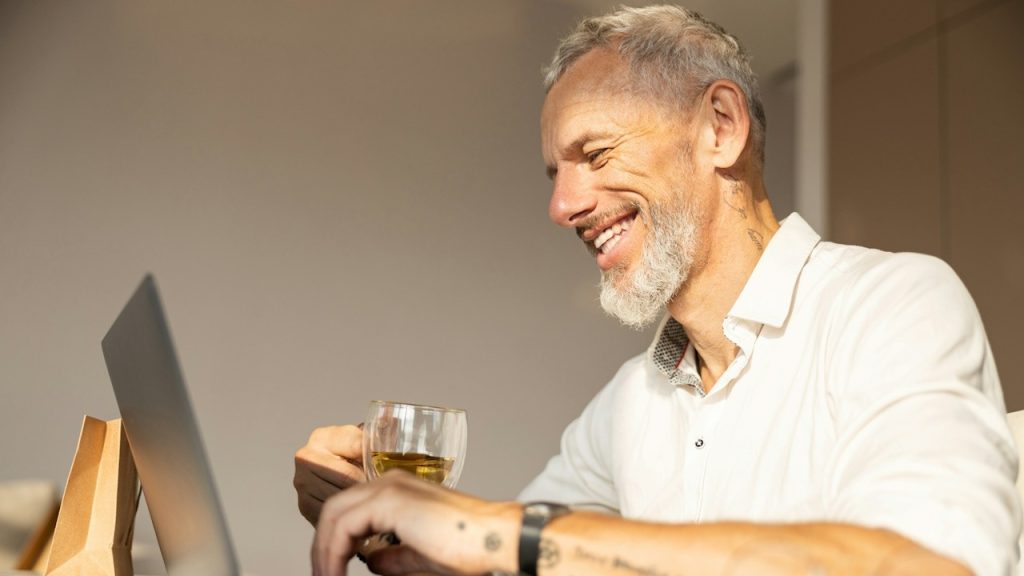 An entrepreneur is pressing the keys on a laptop keyboard and having a tea in a sunlit room.