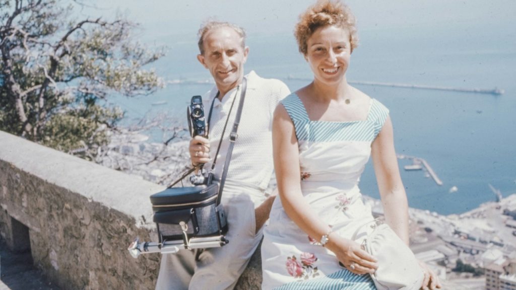 A man holding a huge camera sitting behind a smiling woman as they sit by the sea.