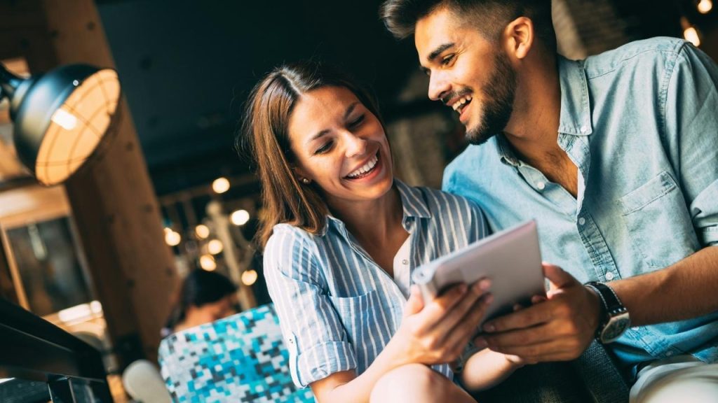 A man and woman laugh while looking at a tablet together in a dimly lit cafe.
