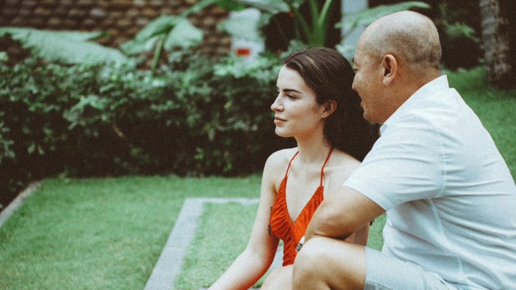 A bald man smiles as he looks at a woman who stares into space while they sit on a lawn.